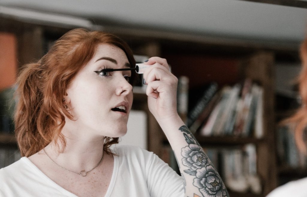 A woman with red hair applies mascara to her eyelashes.