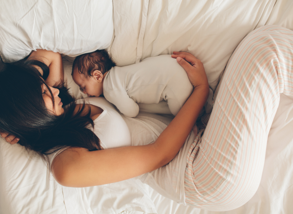 A young woman with long dark hair wearing white pyjamas lays in a bed with a small baby next to her.