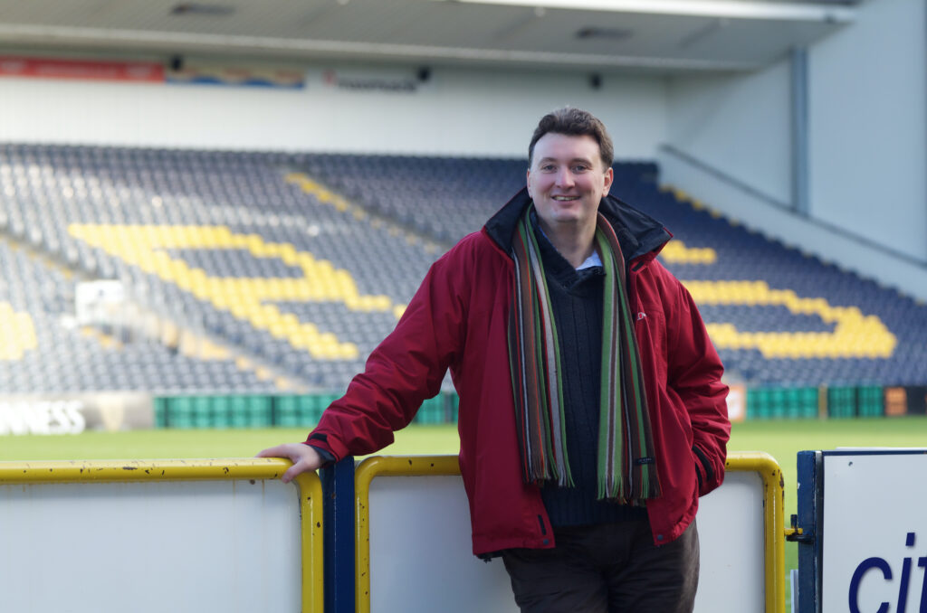 ncmh director Ian Jones standing in a football stadium in a red coat and stripy scarf