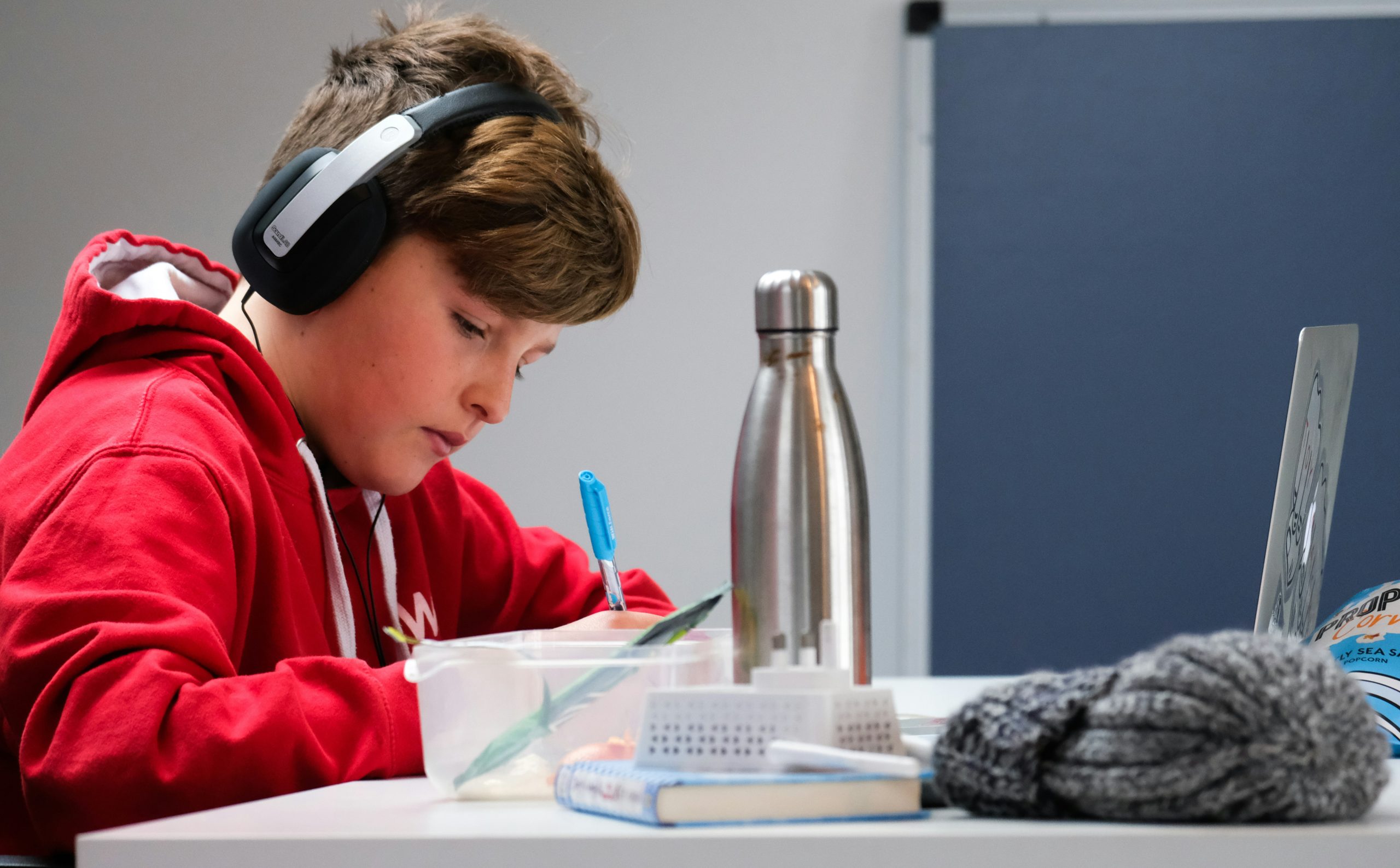Boy in red jumper writes at desk wearing headphones.