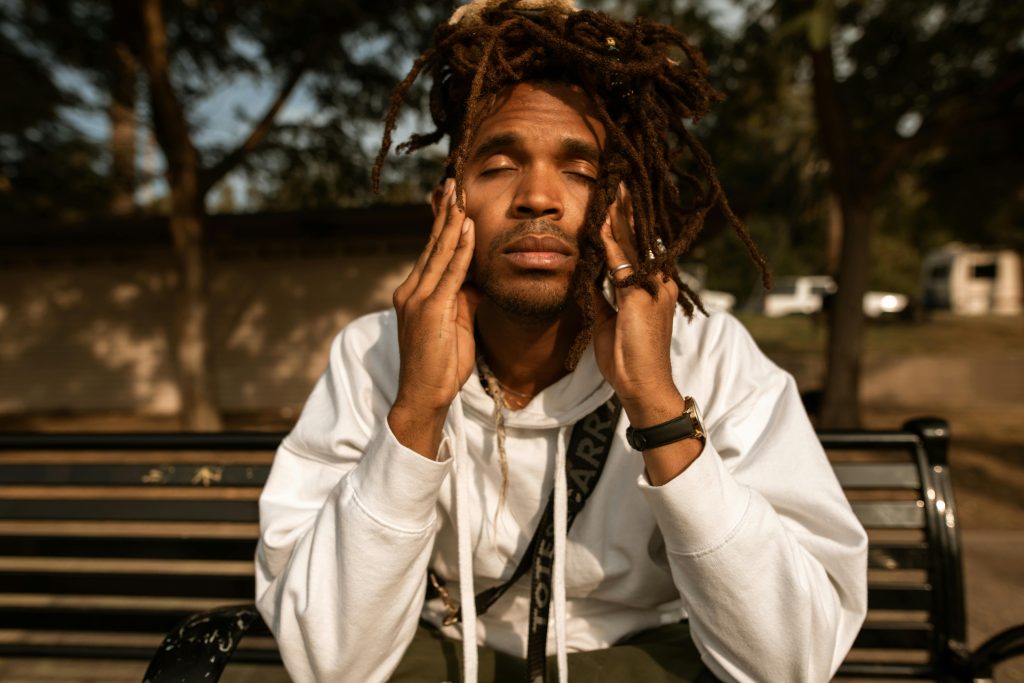 A young black man sits on a bench with his eyes closed and hands touching his temples.