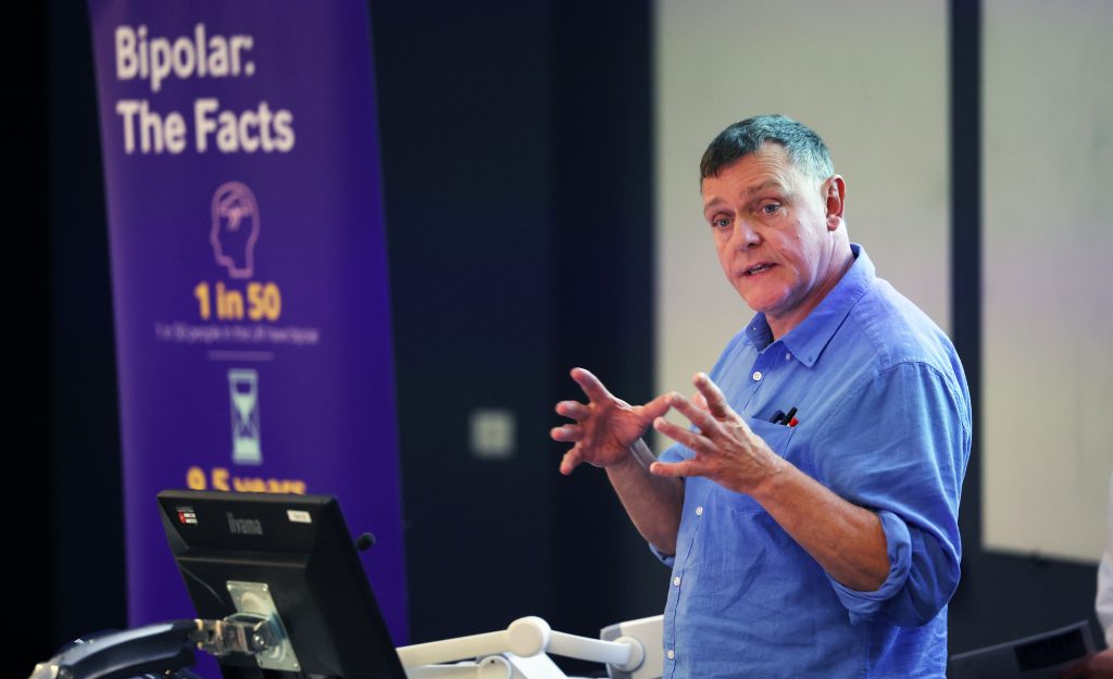John Tredget in a blue shirt speaks at the podium of a Bipolar UK event in 2023.