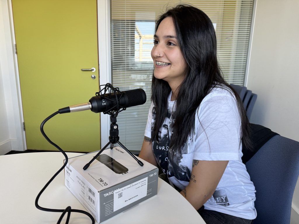 A young woman with long black hair is chatting into a microphone and smiling.