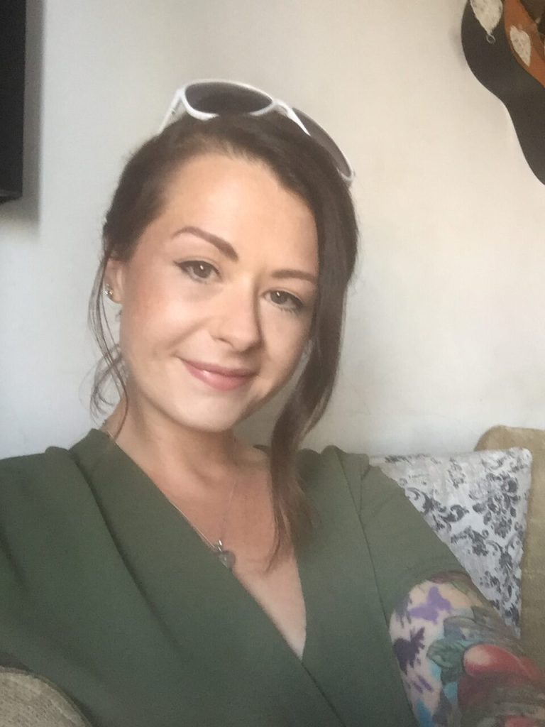 Hayley is a young white woman with long brown hair, she has it up in this photo with white sunglasses on her head. She wears a green v-neck top and is smiling at the camera.