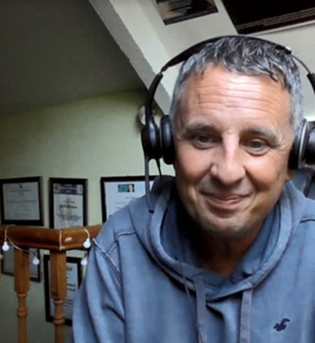 Mark is a white middle aged man with short dark and grey hair. Here he wears a light blue jumper and a set of headphones, and he is smiling at the camera.