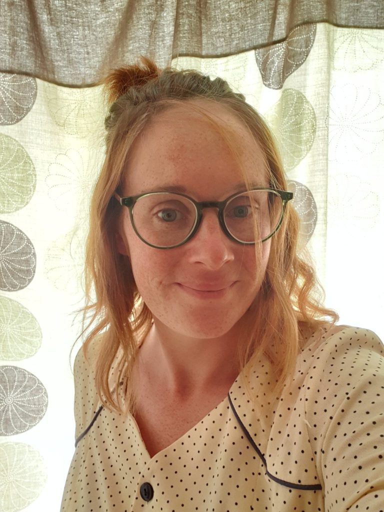 Becci is a young white woman with ginger hair, half tied up on her head and she wears round black-rimmed glasses. She is smiling at the camera and wears a white top with little black dots.
