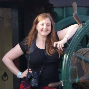 Jayne is a young white woman with long brown hair and she is wearing a black t-shirt with a camera on a strap around her neck. She's leaning on a ship's helm with the other arm on her waist.