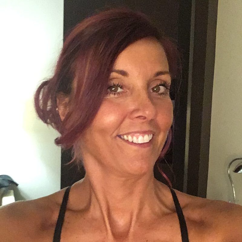 Liz is smiling at the camera and can be seen from the shoulders up in a black halter neck top and her purple brown hair in a loose knot.