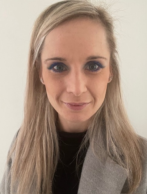 Steph is a young white woman with light blonde hair. She is smiling at the camera wearing a black top and grey scarf.