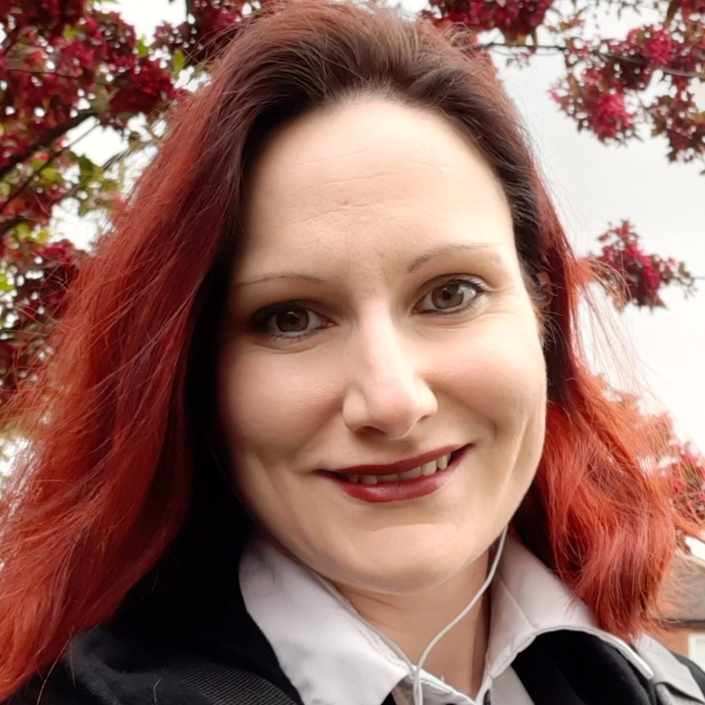 Victoria is a young white woman with dyed red hair and she is smiling at the camera.