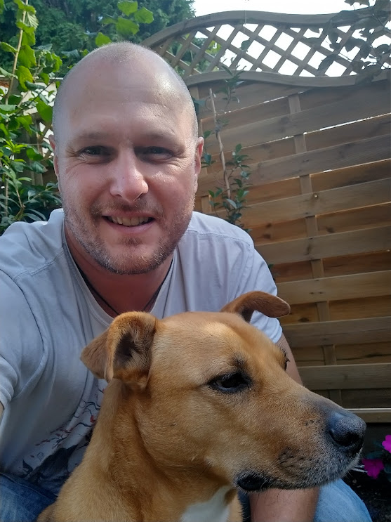 A photo of Champion Steve, he is a middle aged white man with a shaved head and is smiling at the camera, he has a light brown short-haired dog sitting in front of him.