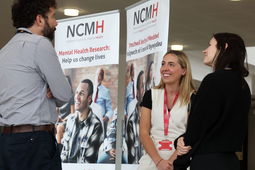 Three people, a brown haired bearded man and two young women, one blonde and one brunette, stand speaking in front of NCMH banners.