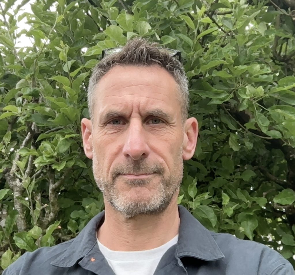 An image of Professor Simon Dymond, Director of the GREAT Centre who is stood in front of a tree. He is a white man with short hair, stubble and is wearing a grey shirt.