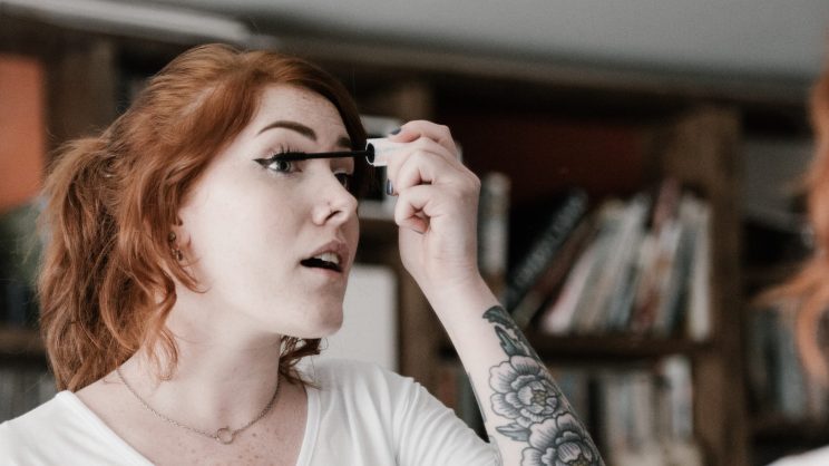 A woman with red hair applies mascara to her eyelashes.