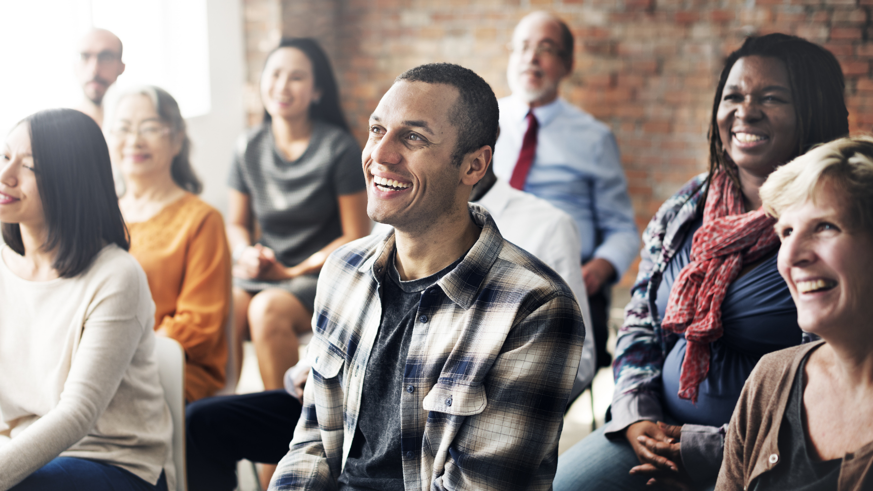 A diverse group of people of different ages and ethnicities sitting in rows with the focus on a smiling young man in a plaid shirt.