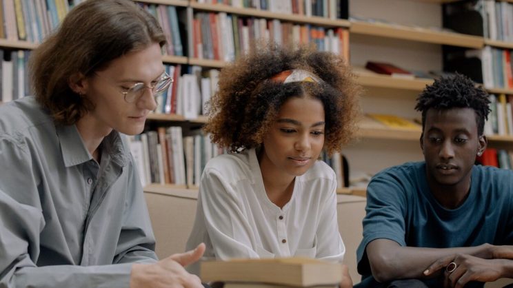 a group of three students sitting at a desk in a library looking at a book together