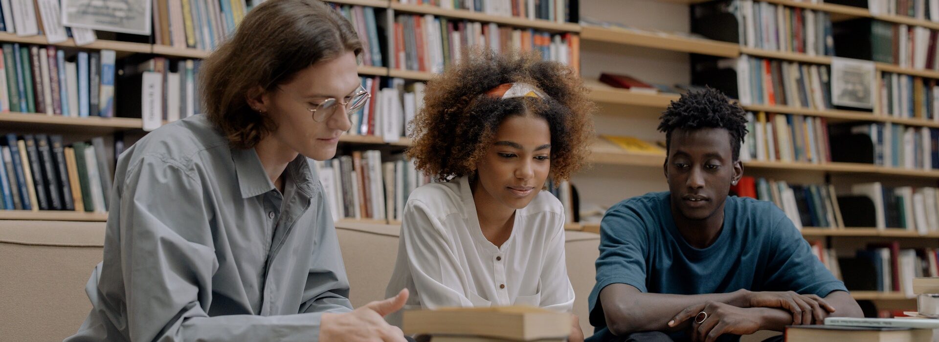 a group of three students sitting at a desk in a library looking at a book together