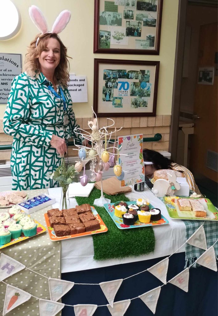 A picture of Barbara wearing a white and green dress and standing next to a bake sale, smiling.