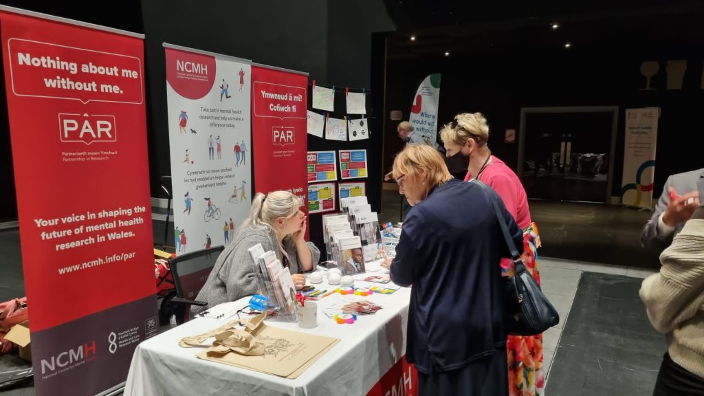 Two people speaking with a person at a conference stand and the banner behind reads PAR which stands for Partnership in Research at NCMH