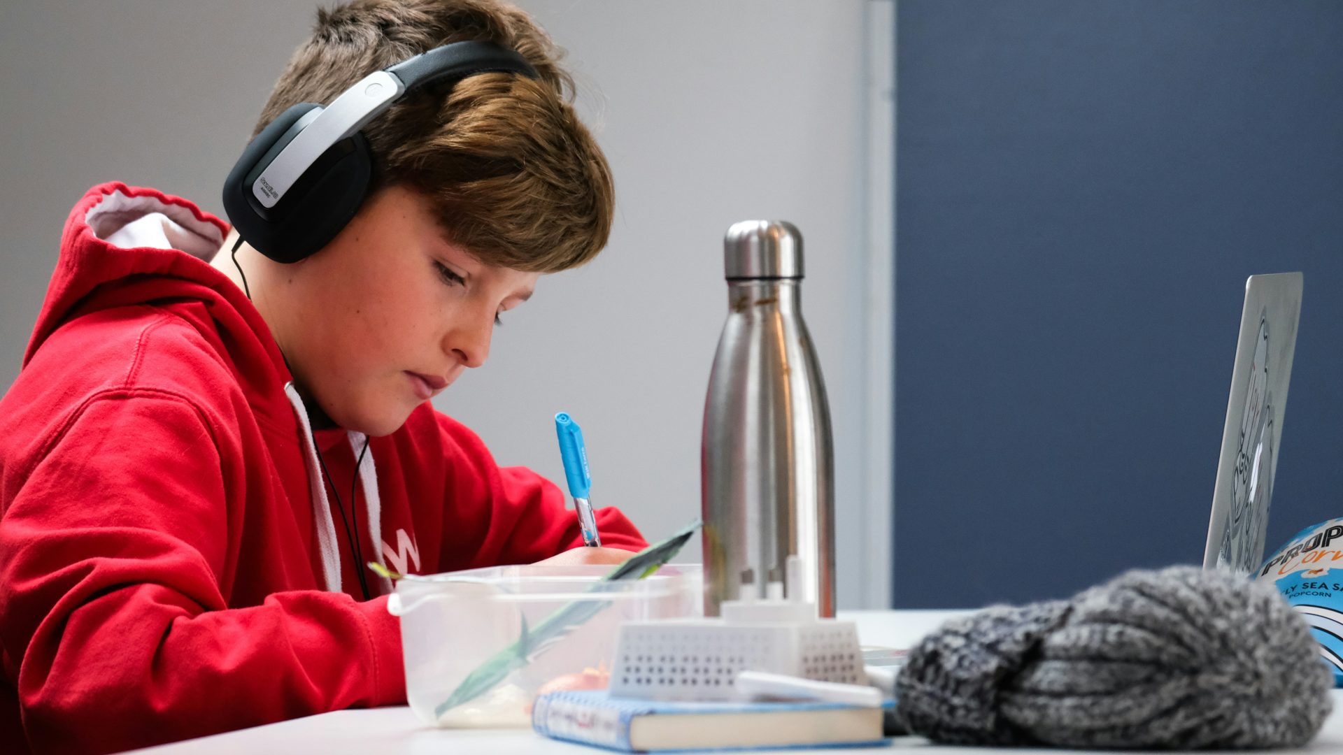 Boy in red jumper writes at desk wearing headphones.