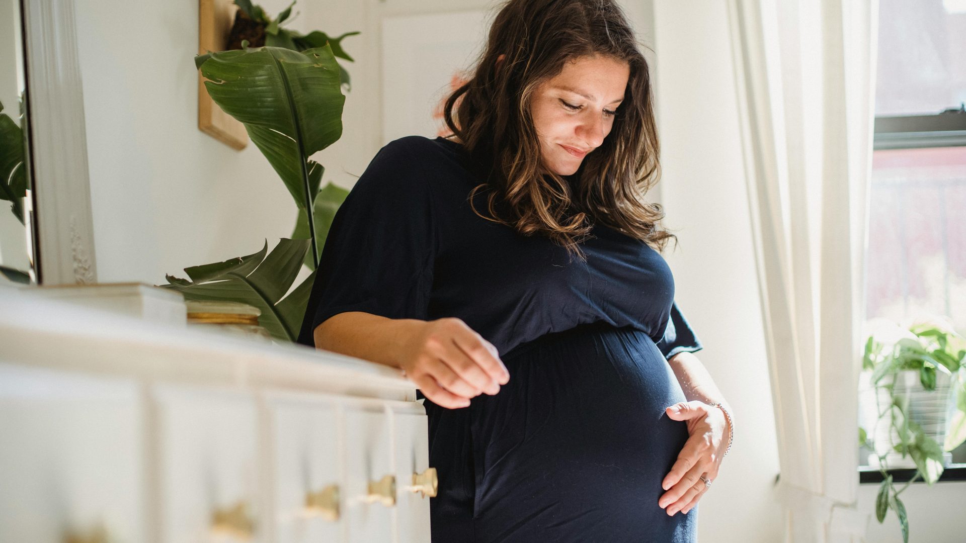 A pregnant woman with brown hair in a navy dress leans on a counter holding her belly.
