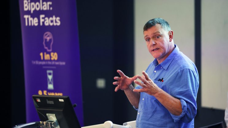 John Tredget in a blue shirt speaks at the podium of a Bipolar UK event in 2023.