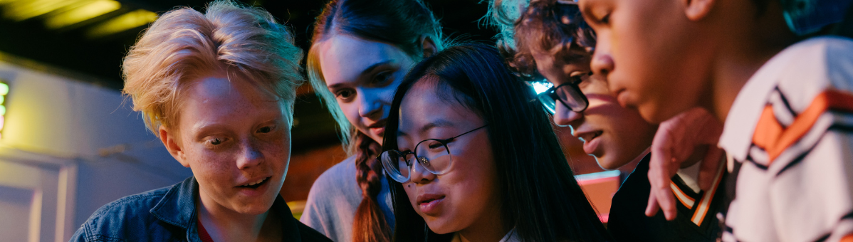 A diverse group of five young teenagers crowd around a magazine. A young Asian girl is in the foreground holding the magazine. She has long dark hair and is wearing glasses and a white denim jacket.