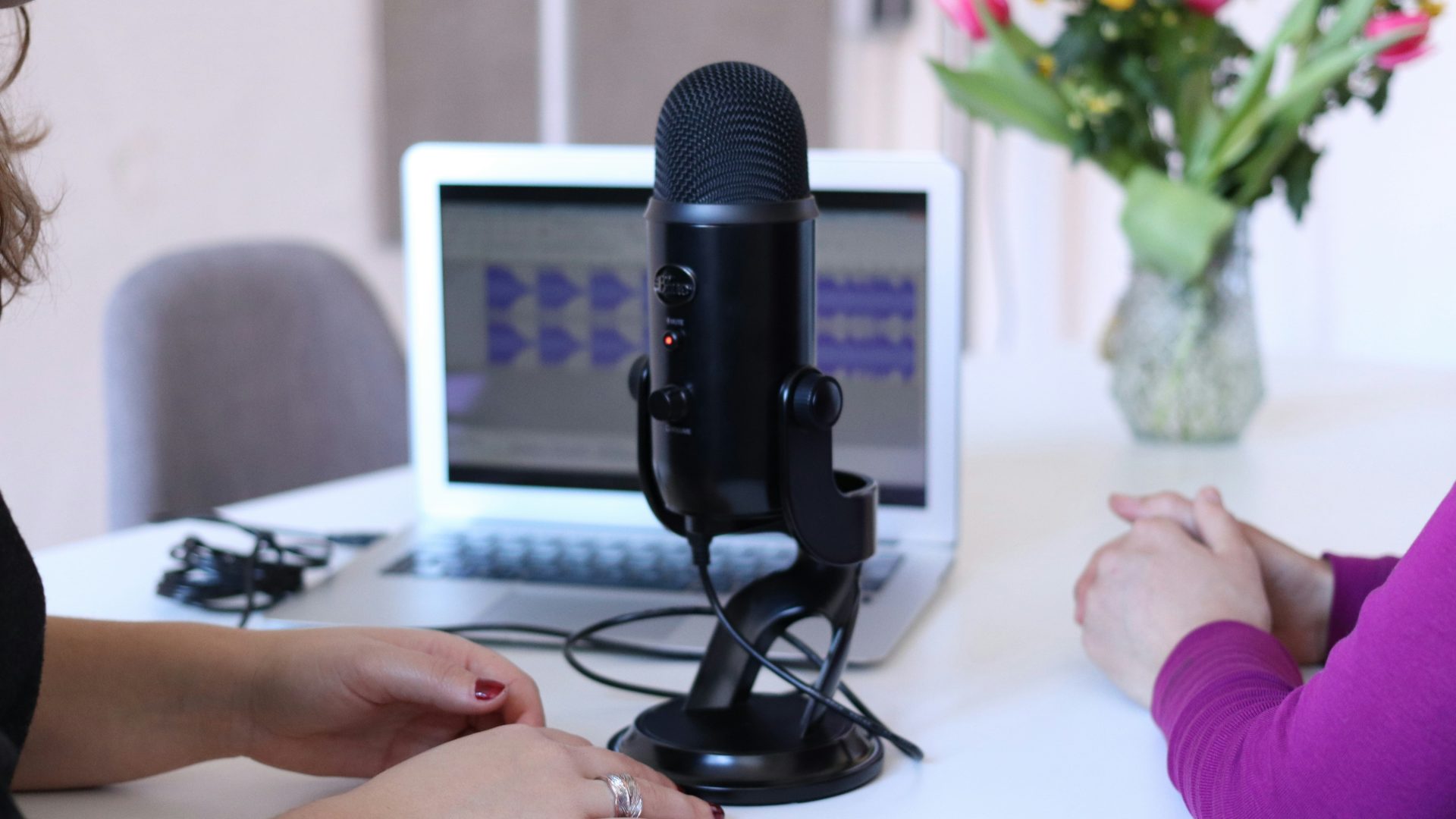 Two women sit in front of a podcast microphone and an open laptop.