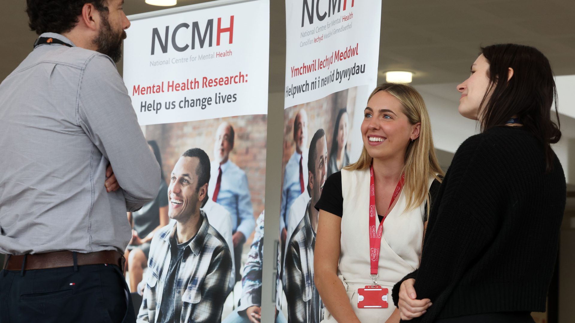 Three people, a brown haired bearded man and two young women, one blonde and one brunette, stand speaking in front of NCMH banners.