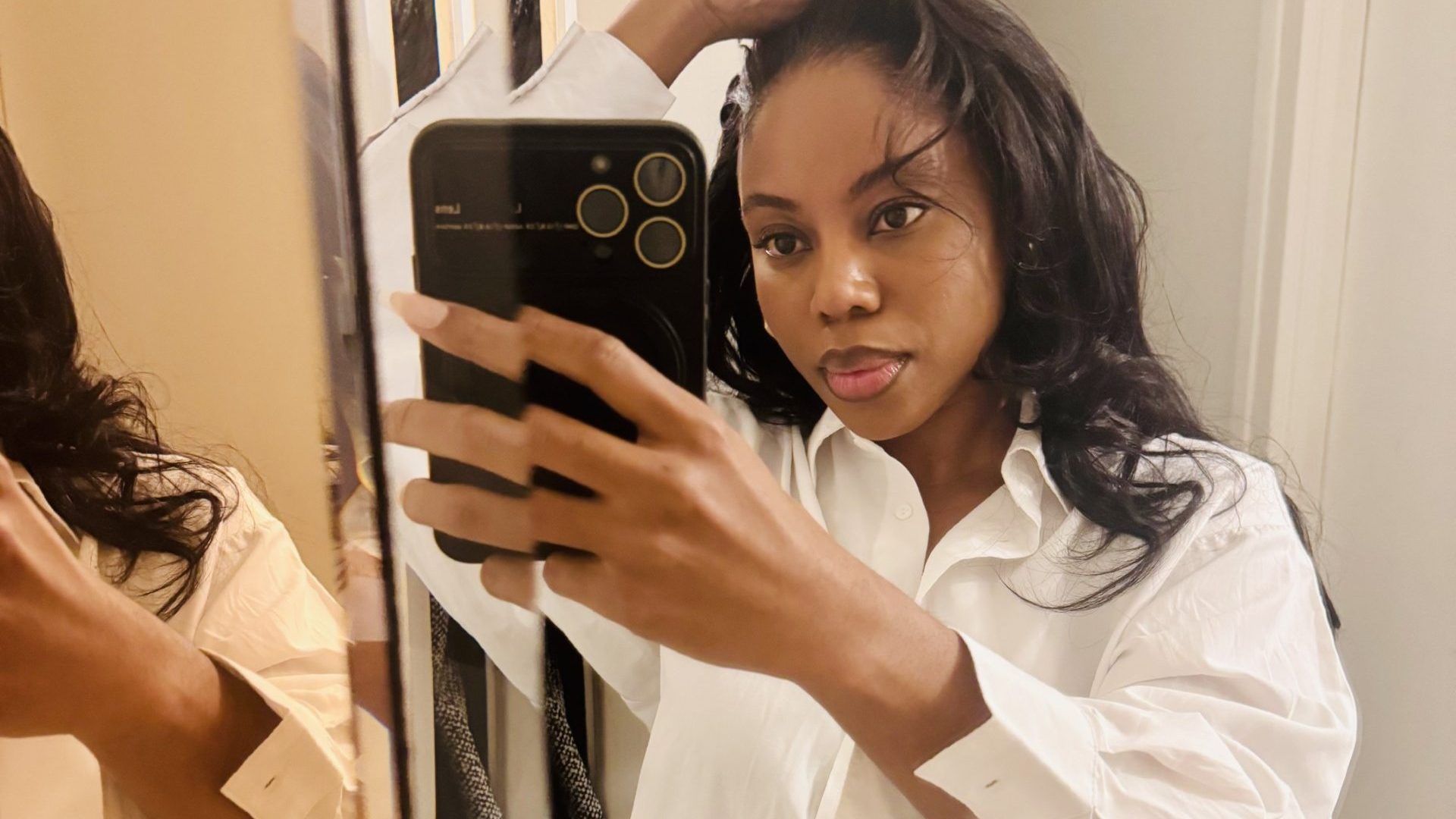 An image of Sandylee who is taking a picture of herself in the mirror. She has dark brown hair and wears a white shirt.